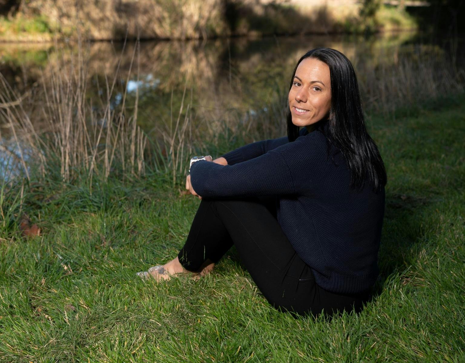 Minda Murray, a woman with long dark hair and wearing dark clothing, sits on the grass next to a pond.
