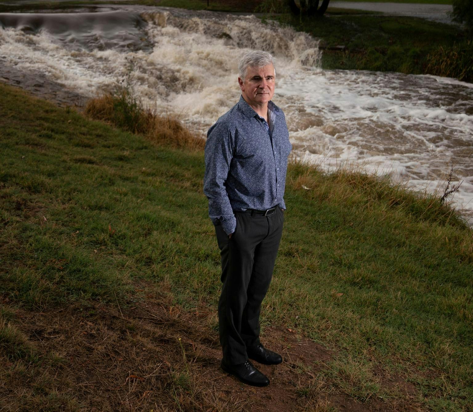 Mark Howden is wearing a blue collared shirt and black pants. He is standing on a grass bank and behind him is a river way. with frothy white-brown water flowing through
