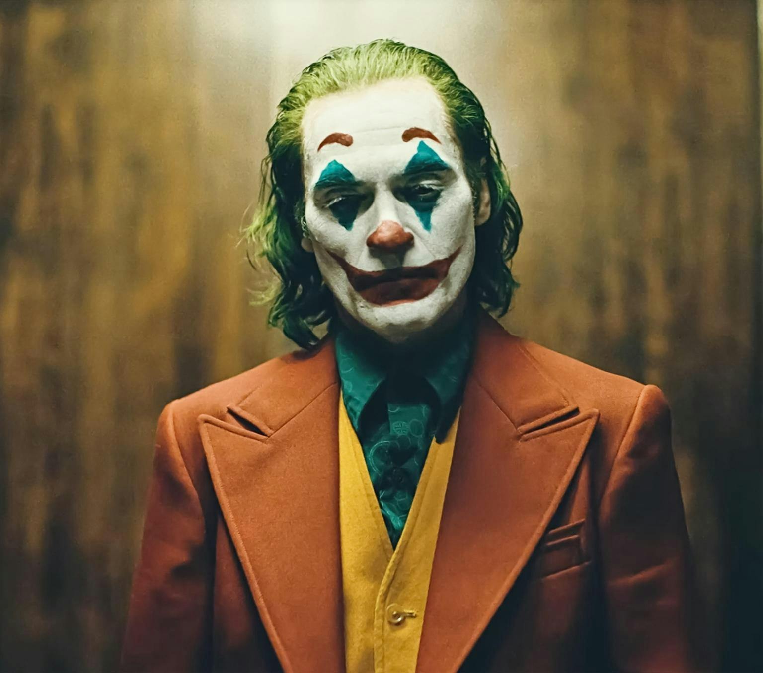 Joaquin Phoenix as the Joker, with his face painted in clown make up including a big red smile that extends onto his cheeks, stands in an orange suit, yellow vest and green shirt and looks downcast.