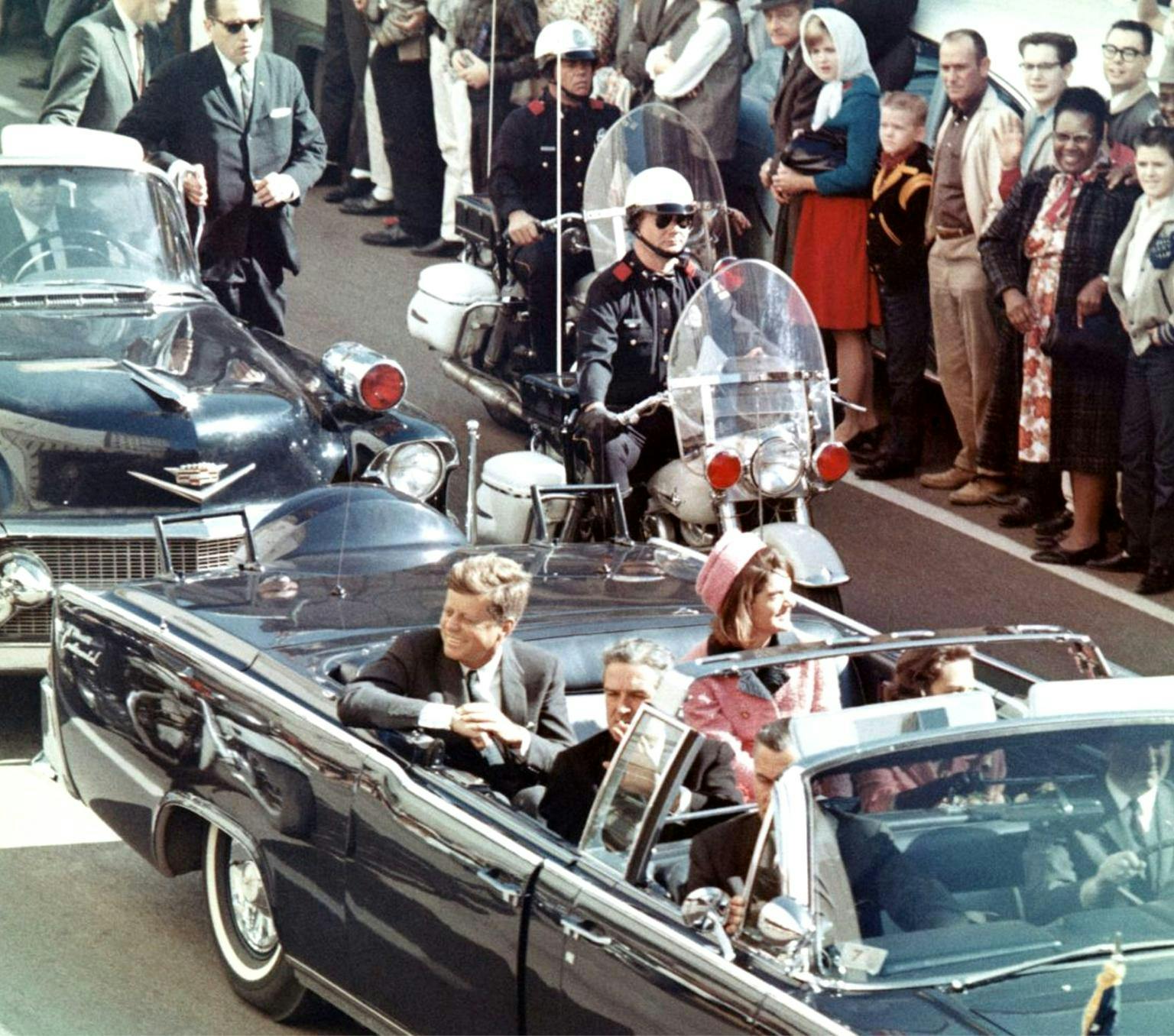 President John F Kennedy sits in the back seat of a convertible limousine. A policeman on a motorbike follows the car. The street is lined with people.