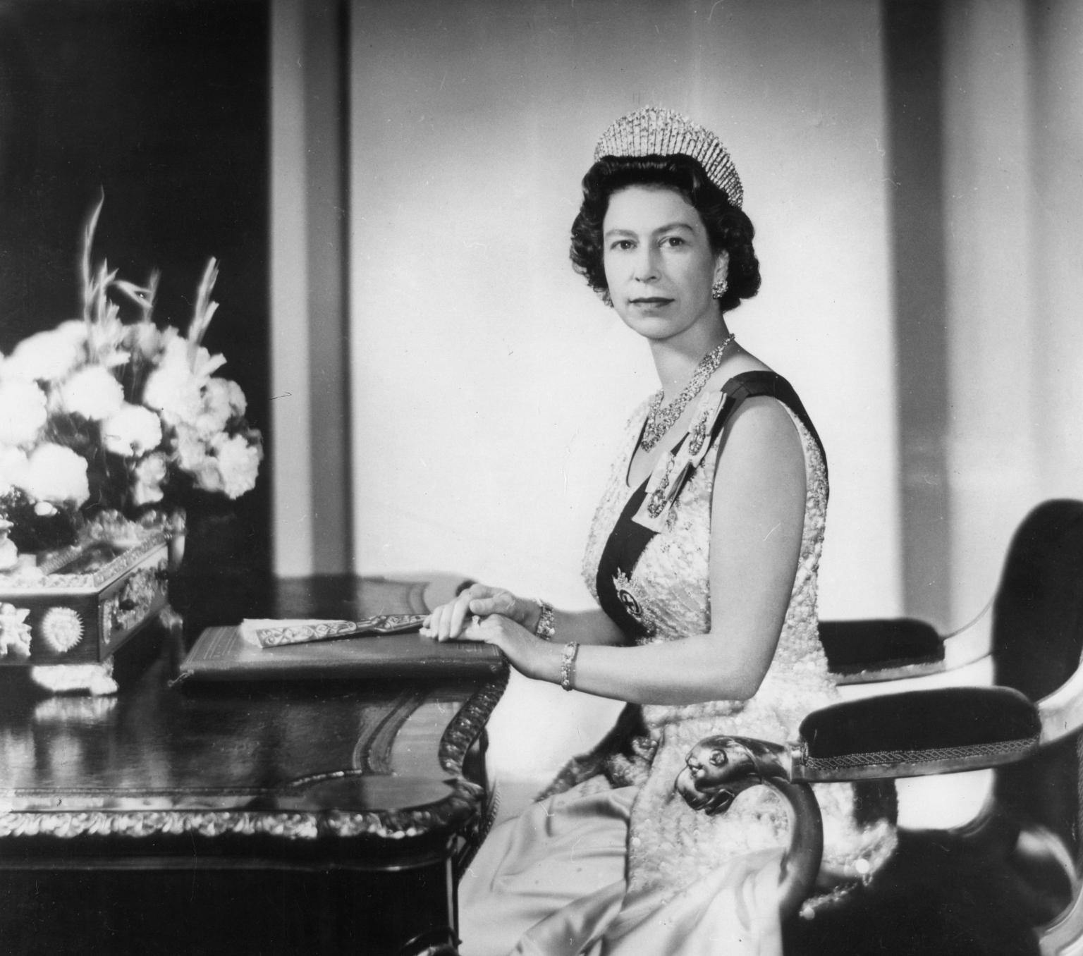 Queen Elizabeth, wearing a tiara and sash, sits at a table and looks at the camera.