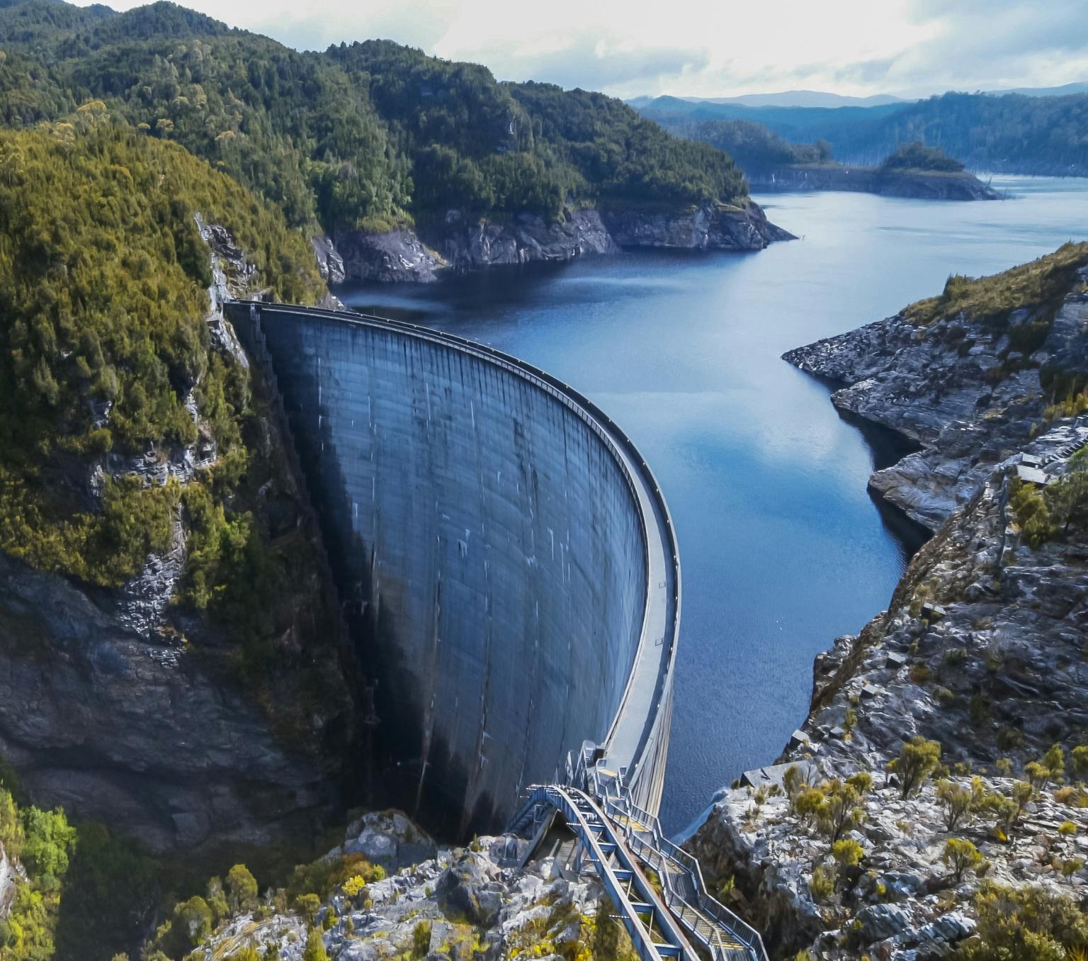 A large concrete dam wall, with a large body of water on one side, surrounded by mountains and forest.