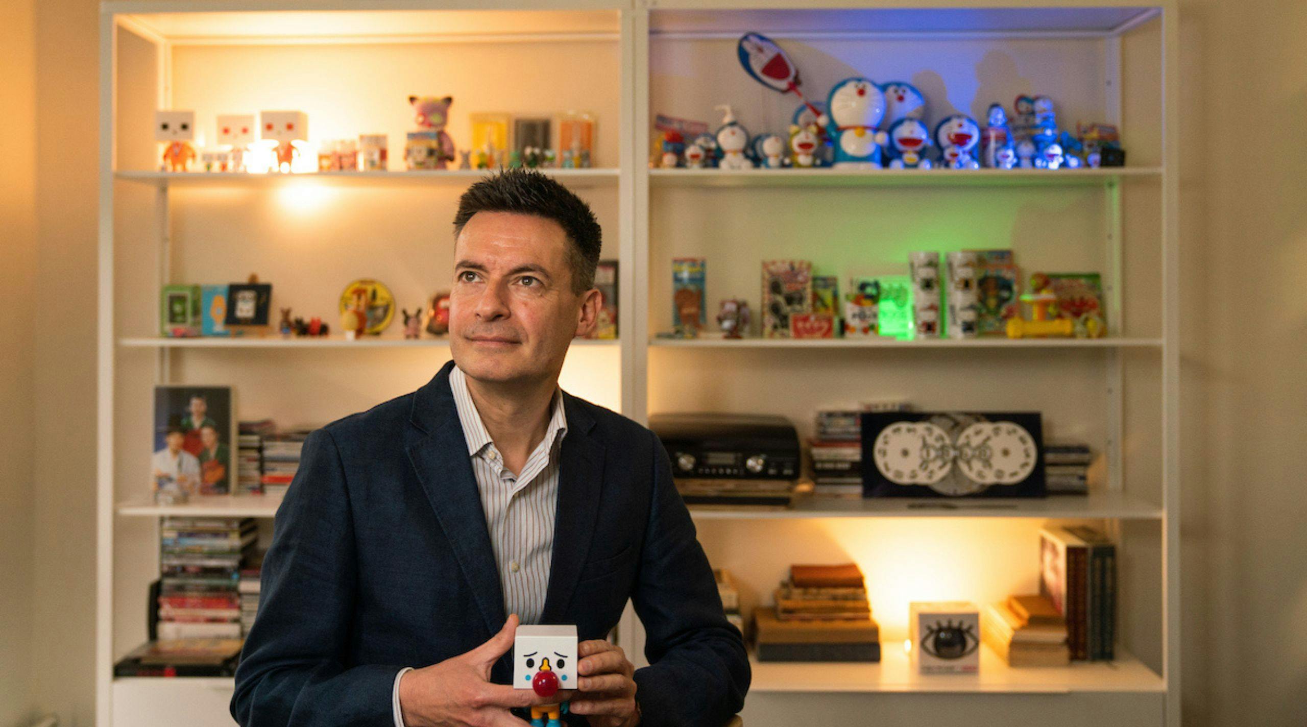 Roald Maliangkay in his office, he is holding a toy that looks like tofu. Behind him are shelves filled with designer toys, with many different versions of the blue robot cat Doraemon.