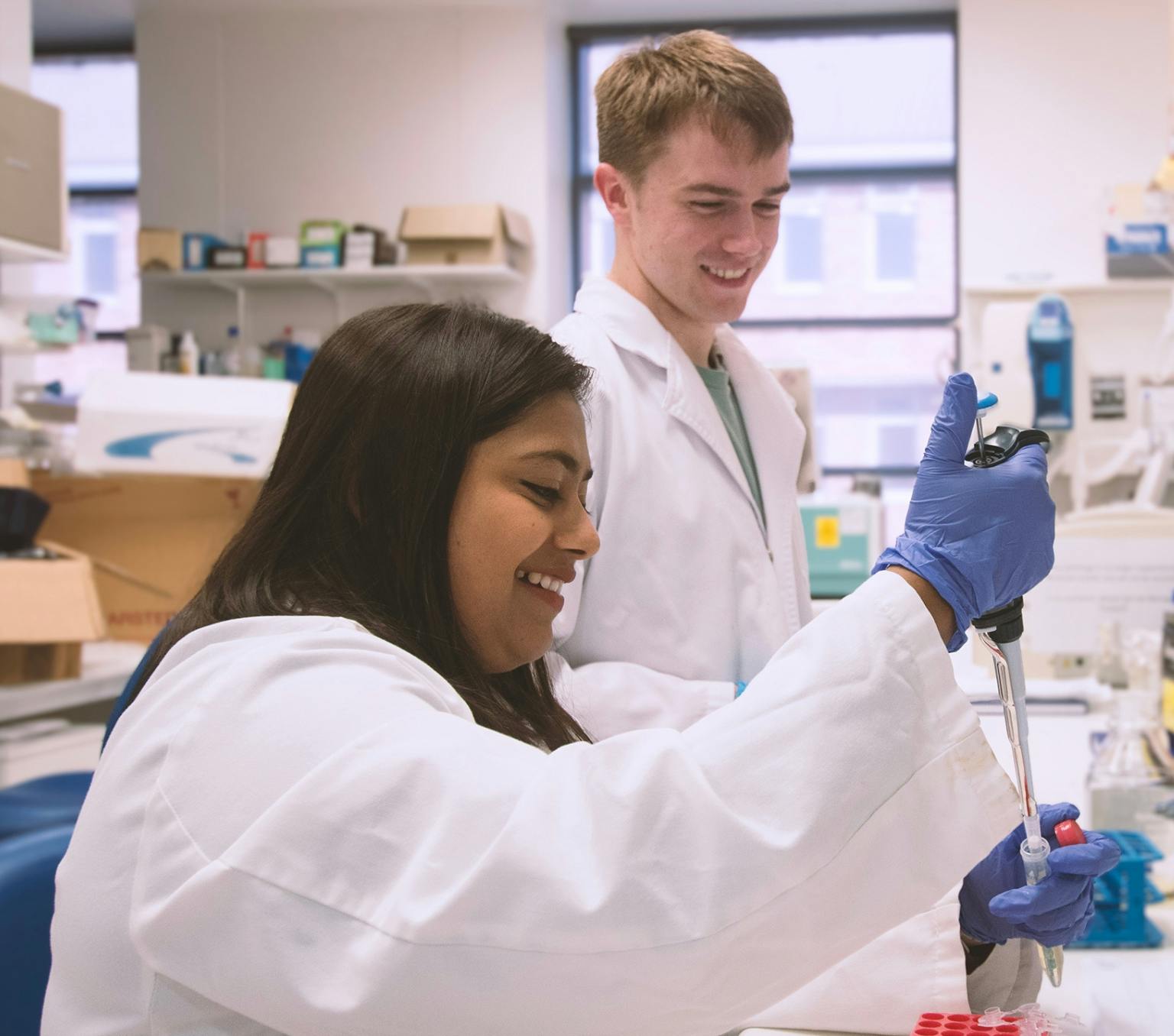 Dr Anukriti Mathur and Callum Kay in a science lab. They are both wearing white lab coats. Dr Mathur is injecting an unknown substance into a test tube.